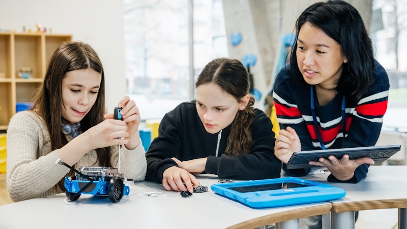 Two female students working together to build a blue robot car. A teacher is observing to the side.