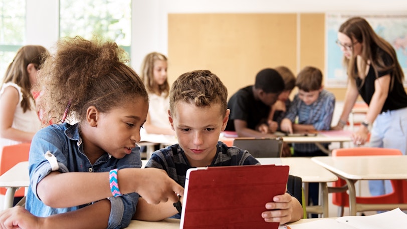 Two young students sitting together in a classroom and looking at a tablet screen with a red protective cover on it.